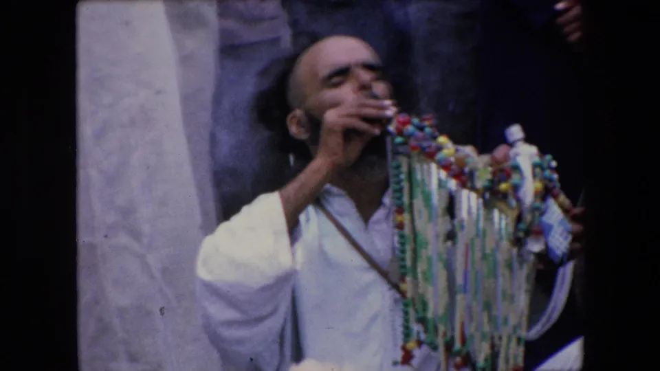 1971 Algeria home-movie frame showing a seated man in white clothing smoking beside a large strand of colorful beads, with partial figures in the background.