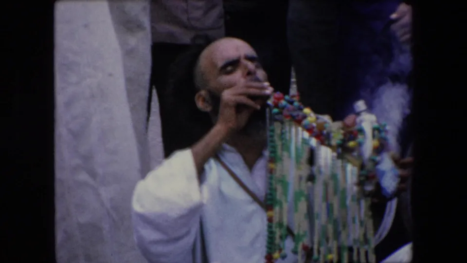 1971 Algeria frame of a man in white clothing exhaling smoke next to colorful hanging beads, with partial onlookers and a pale column behind him.