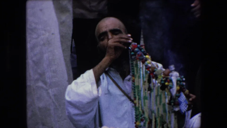 A seated man in a white robe appears to smoke while holding or standing beside colorful bead strands in 1971 Algeria home-movie footage.