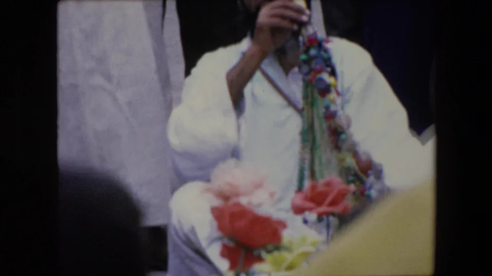 A robed figure is partly obscured while colorful bead strands and soft foreground flowers fill the frame in the 1971 Algeria market scene.