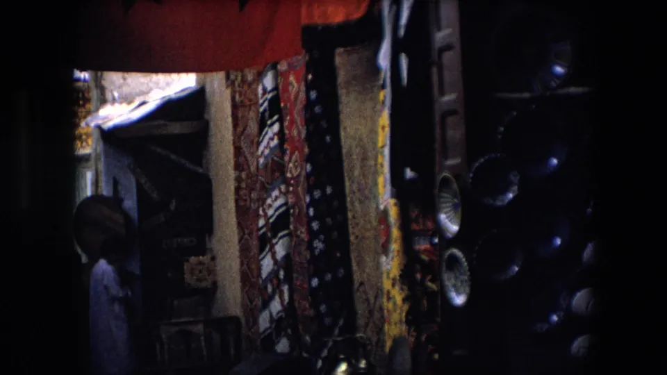 A dim market passage in Algeria shows hanging patterned textiles, shopfront details, stacked speakers, and a person standing in shadow.