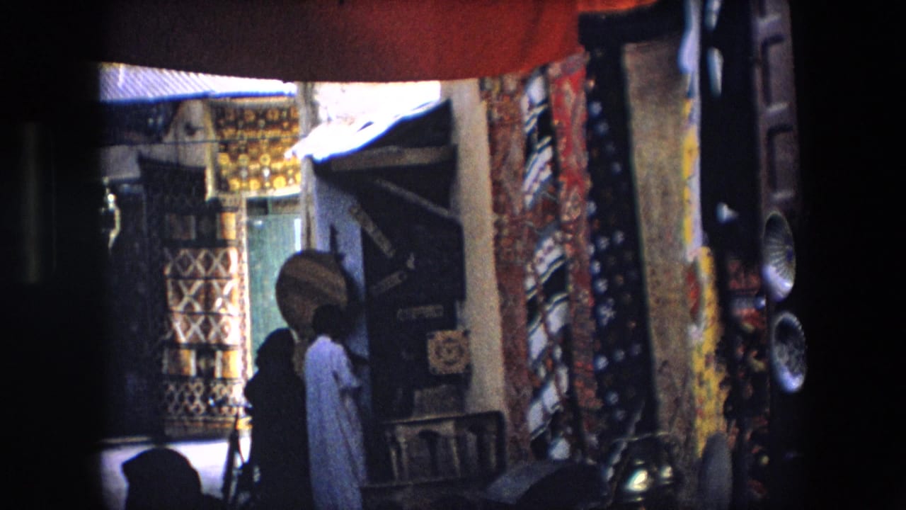 Shaded market alley with hanging rugs, textiles, and pedestrians near a stall.