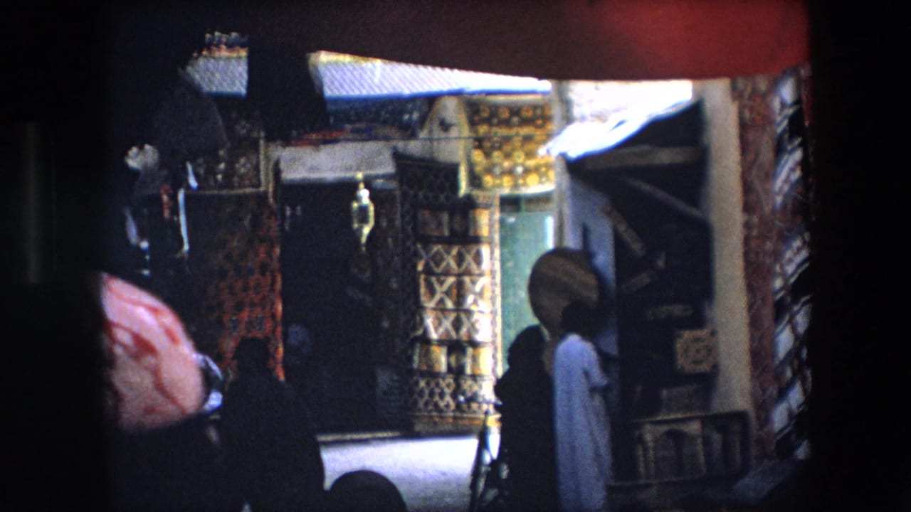 1971 Algeria market frame with patterned rugs and fabric displays partly obscured by a dark foreground figure and shadowed camera framing.