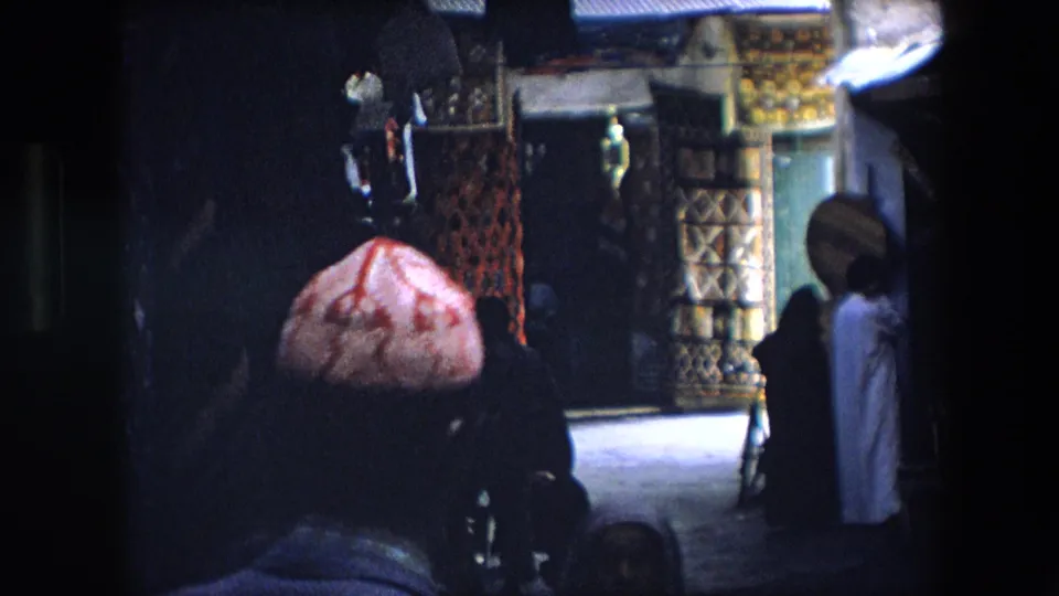 1971 Algeria 8mm frame from a low, shadowed angle showing rugs and textiles along a market passage with pedestrians near the stall.