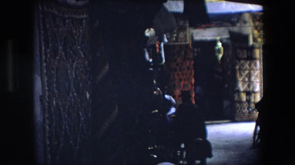 Final 1971 Algeria market frame showing a dim, partially blocked view of hanging rugs and textiles with people moving near the stall frontage.