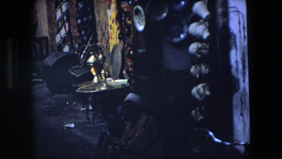1971 Algeria 8mm frame showing a close view of a market stall with hanging goods, patterned textiles, metalware, and ceramic or household items.