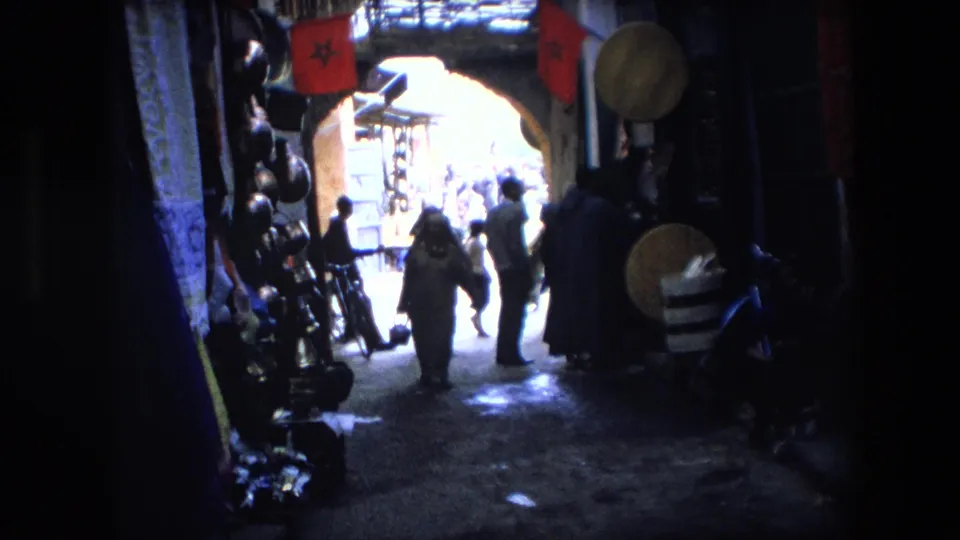 1971 Algeria 8mm frame showing several people walking through a shaded market alley toward a bright street opening with goods displayed on both sides.