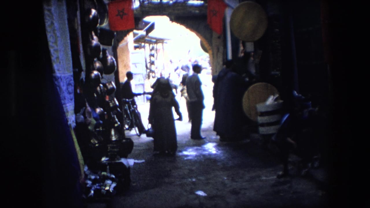 1971 8mm footage shows pedestrians, including women in draped clothing, moving through a shadowed North African marketplace passage with red flags and hanging goods visible near a bright archway.