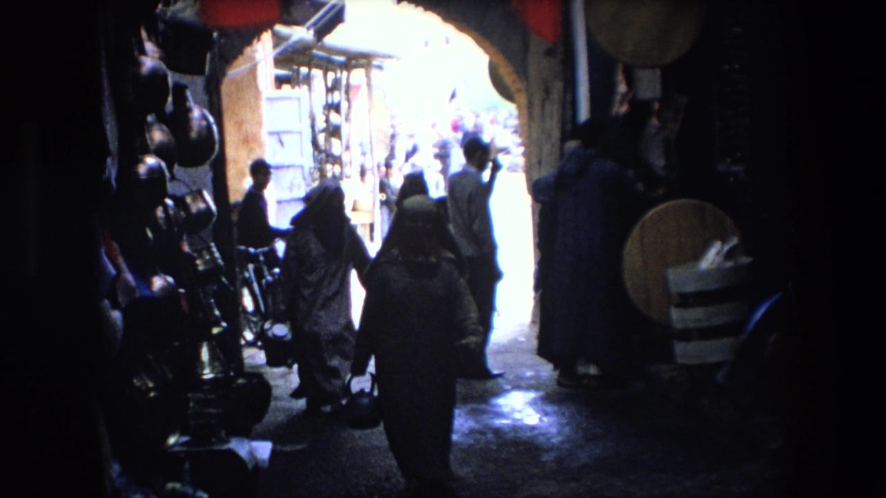 Several people walk through a dark passage lined with goods and red flags near a bright archway.