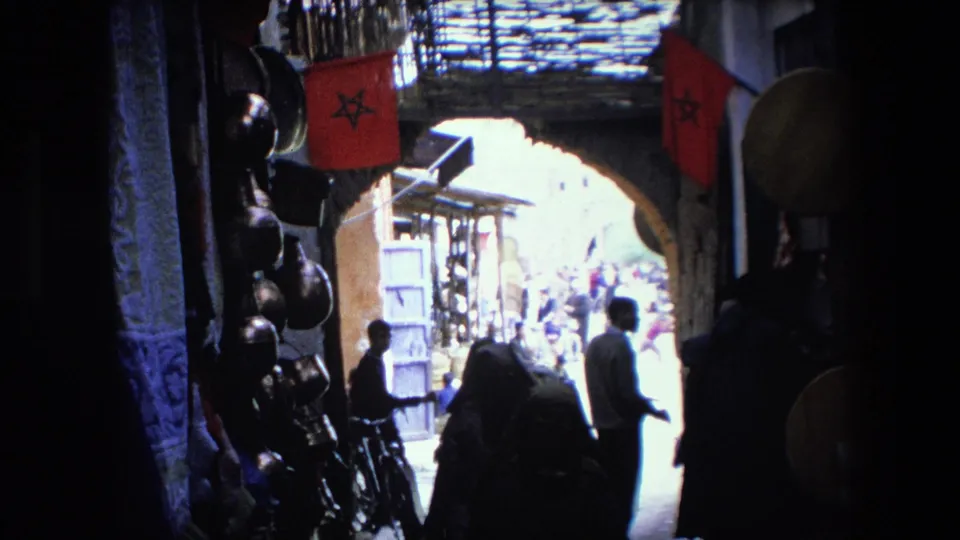 The frame shows a shadowed 1971 8mm market passage with a woman in draped clothing in the foreground, other pedestrians near the bright exit, hanging goods, and red flags overhead.