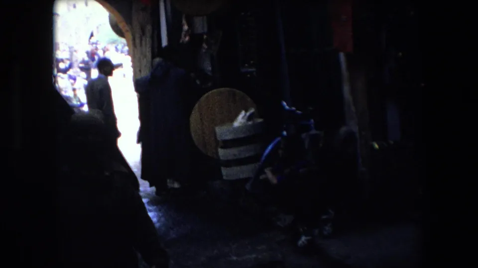 This final frame from the 1971 8mm clip shows a silhouetted pedestrian near the bright end of a marketplace alley, with hanging goods, stacked items, and red flags partly visible in deep shadow.