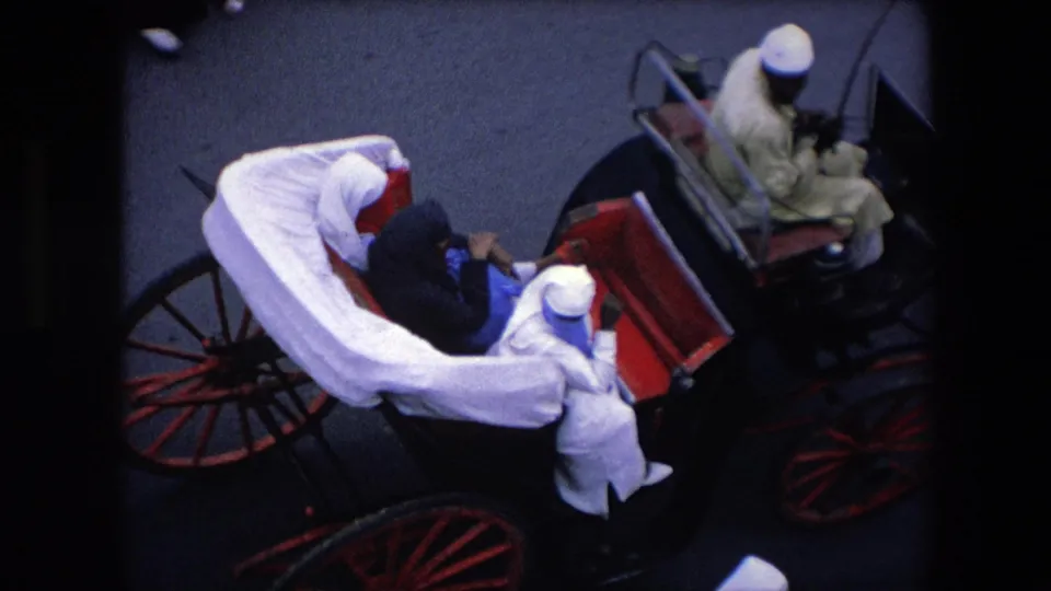 1971 Algeria home-movie frame with passengers seated in a white-draped horse-drawn carriage, red carriage panels, large wooden wheels, and another carriage visible to the side.