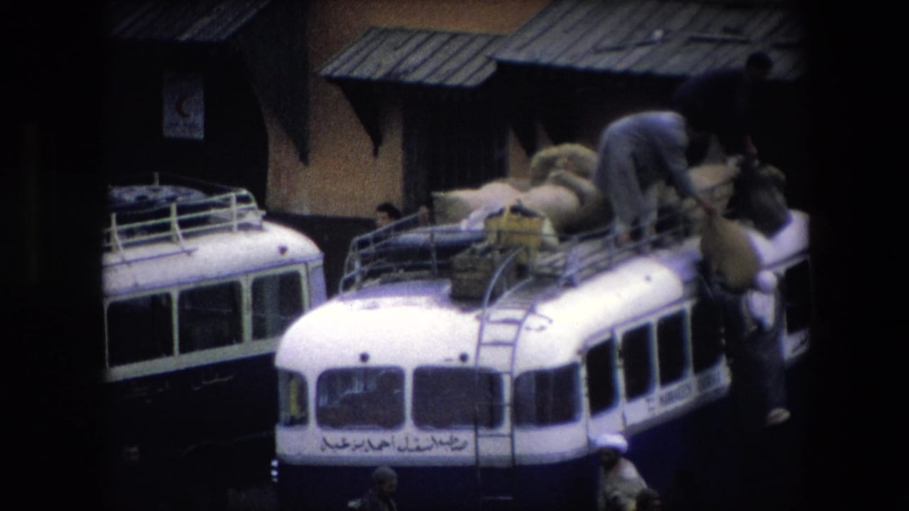 In 1971 Algeria, men work around a white public bus with Arabic lettering as large sacks are arranged on its roof rack beside a building with metal awnings.