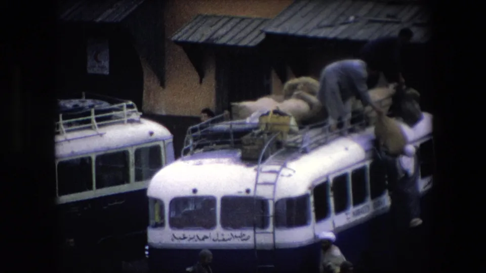 In 1971 Algeria, men work around a white public bus with Arabic lettering as large sacks are arranged on its roof rack beside a building with metal awnings.