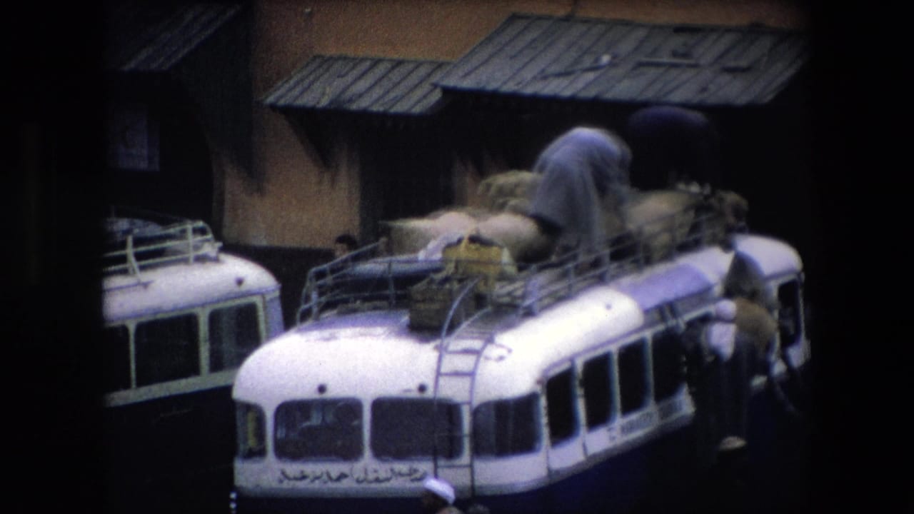 A white public bus in Algeria is shown with a man bent over its roof rack, arranging bulky sacks while another figure stands beside the bus near the building.