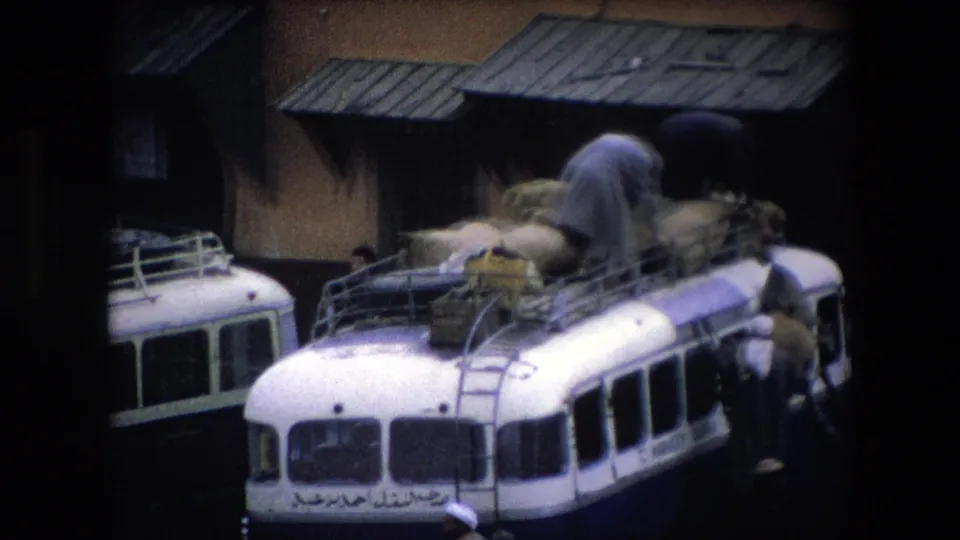 A white public bus in Algeria is shown with a man bent over its roof rack, arranging bulky sacks while another figure stands beside the bus near the building.