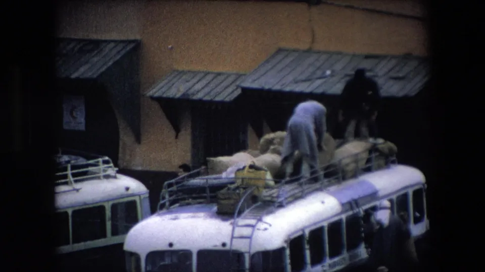 The home-movie frame shows men securing large sacks on the roof of a white bus in 1971 Algeria, with another bus partly visible at left and a wall with corrugated awnings behind them.
