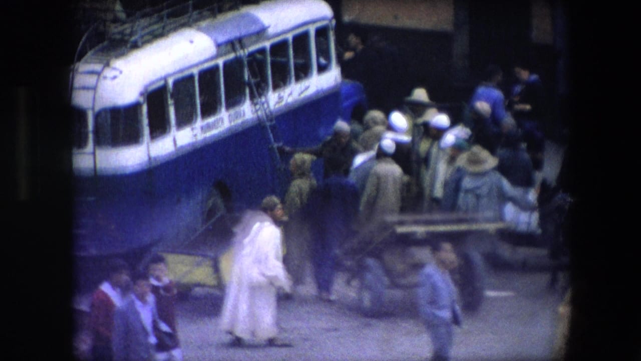 1971 Algeria home-movie frame showing a blue and white bus near a crowd of pedestrians on a street, seen with soft 8mm color and grain.