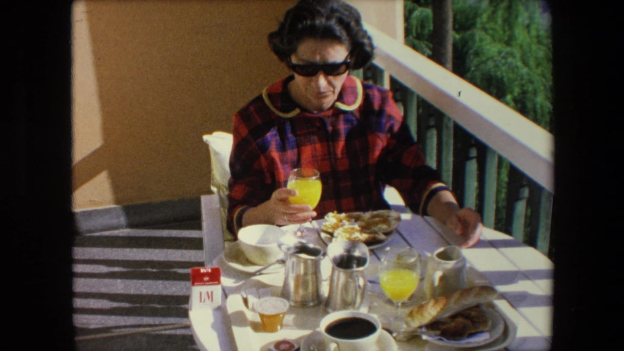Woman in dark sunglasses and a red and blue plaid top sits at a sunlit balcony breakfast table in Algeria in 1971, holding a glass of orange juice beside coffee, bread, and dishes.