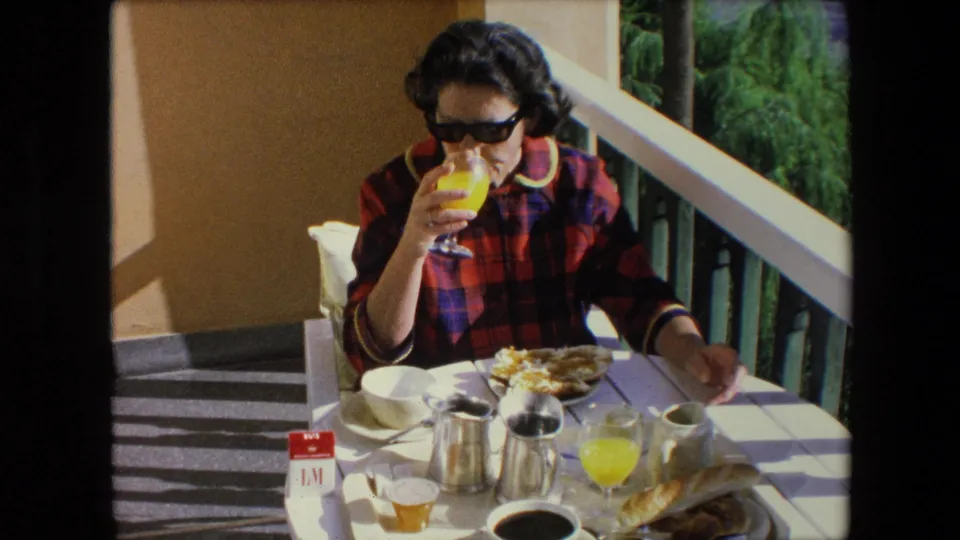 1971 Algeria balcony breakfast frame with the woman in sunglasses raising orange juice while plates, coffee service, bread, and a cup remain arranged on the sunlit table.