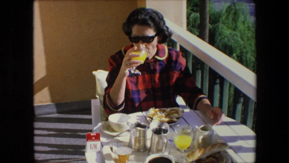 Woman wearing sunglasses drinks orange juice at a balcony breakfast table in 1971 Algeria, with coffee, bread, dishes, railing, and greenery visible around her.