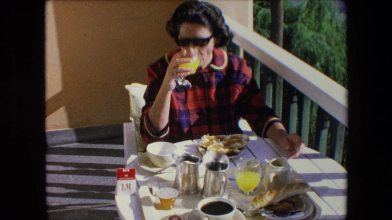 In 1971 Algeria home-movie footage, a woman in dark sunglasses and a red plaid shirt drinks orange juice at a sunlit balcony table set with coffee, bread, and cigarettes.