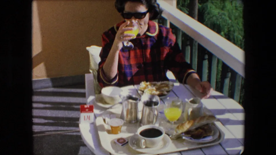 A woman seated outdoors in Algeria in 1971 lifts a glass of orange juice beside a white breakfast table with coffee, bread, metal tableware, and a visible cigarette pack.