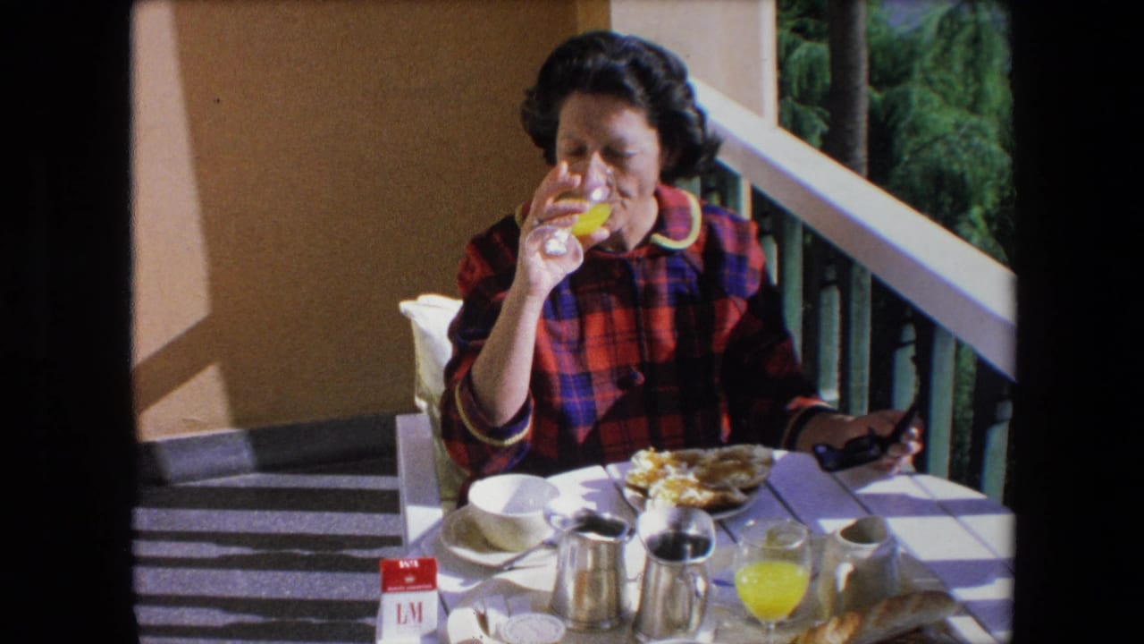 In 1971 Algeria home-movie footage, a brunette woman in a red and blue plaid dress drinks orange juice while seated at a sunlit patio table with coffee service and breakfast dishes.