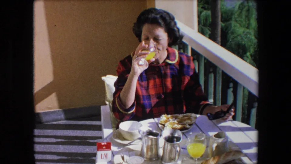 In 1971 Algeria home-movie footage, a brunette woman in a red and blue plaid dress drinks orange juice while seated at a sunlit patio table with coffee service and breakfast dishes.
