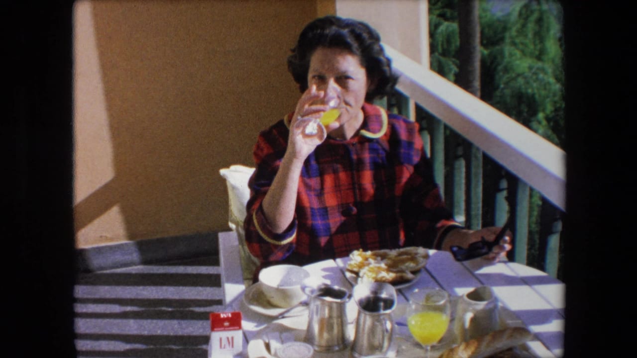 A brunette woman drinks orange juice on a patio in Algeria, with a white table, metal pitchers, coffee cup, breakfast plate, and greenery visible beyond the railing.