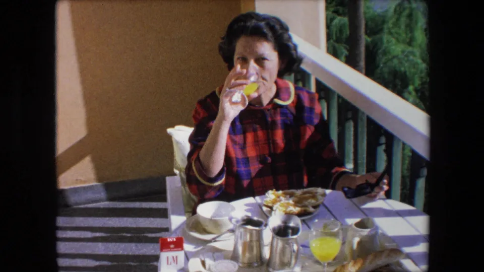 A brunette woman drinks orange juice on a patio in Algeria, with a white table, metal pitchers, coffee cup, breakfast plate, and greenery visible beyond the railing.