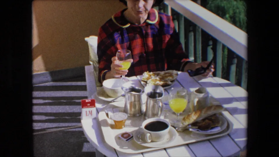 A woman in a red and blue plaid dress sits at a sunlit patio breakfast table in Algeria, holding orange juice beside coffee, bread, dishes, and sunglasses.