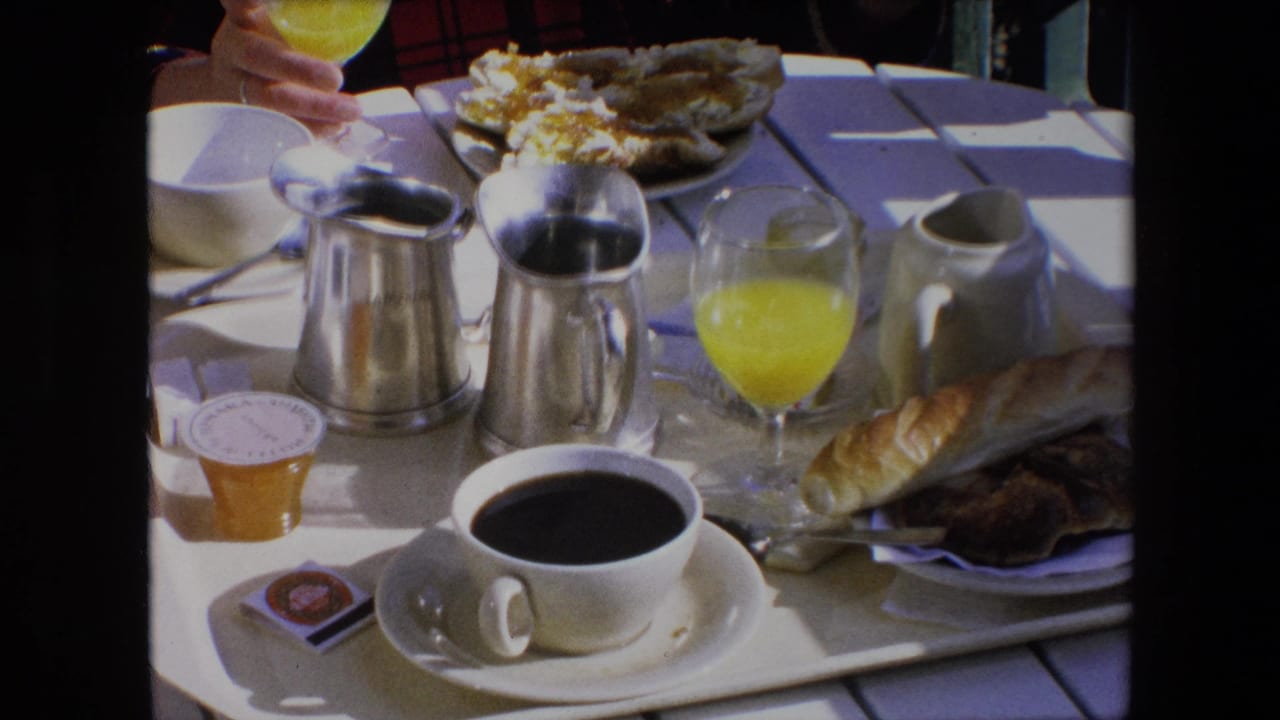 1971 Algeria home-movie frame showing a sunlit outdoor breakfast table with a white coffee cup, orange juice, bread, jam, and silver creamers.