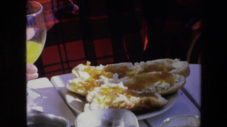 1971 Algeria home-movie close view of bread spread with butter or jam on a plate, with a partially visible orange juice glass and plaid shirt behind it.
