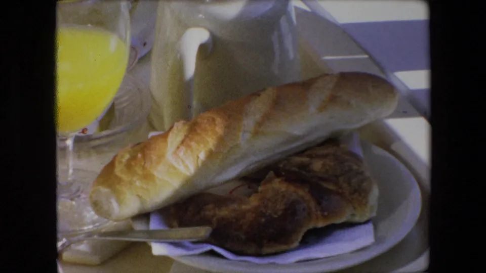 1971 Algeria 8mm frame showing a breakfast plate with a long bread roll, darker pastry, orange juice glass, small knife, and cream-colored pitcher on a tabletop.