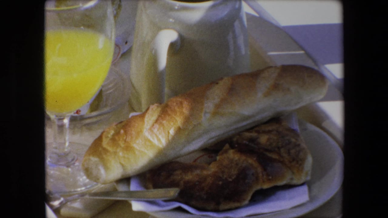 1971 Algeria home-movie frame with a close breakfast table view of bread, pastry, orange juice, a cream-colored pitcher, plate, napkin, and knife.