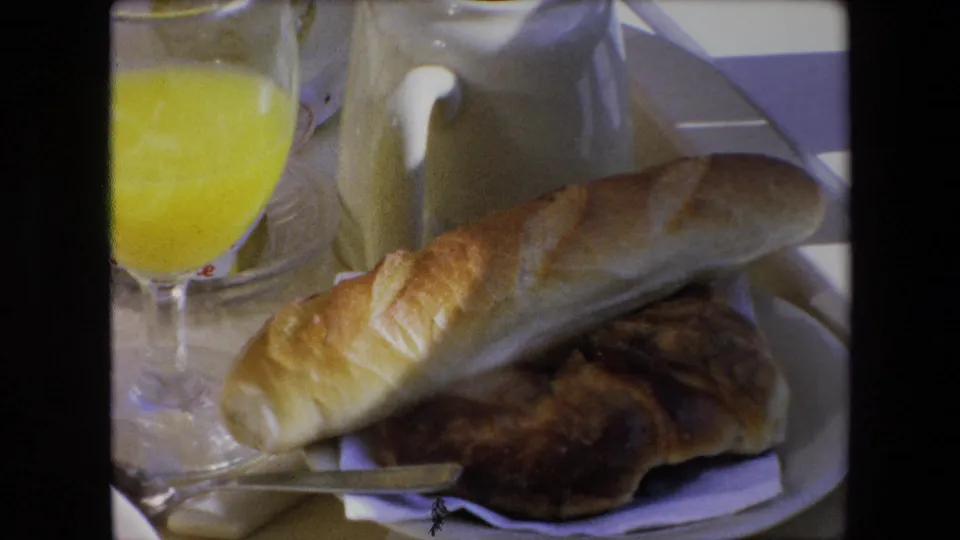 Soft 8mm frame from Algeria in 1971 showing a bread roll and pastry on a white plate with orange juice, a small knife, and a cream-colored pitcher in the background.