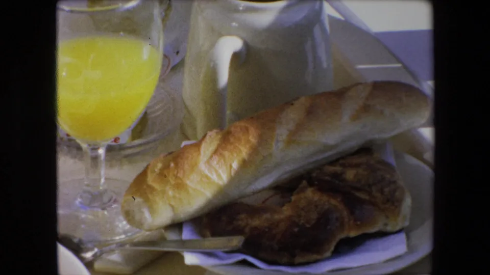 1971 Algeria breakfast close-up on 8mm film, with a long bread roll over a darker pastry, a white plate, folded napkin, small knife, orange juice glass, and pitcher.