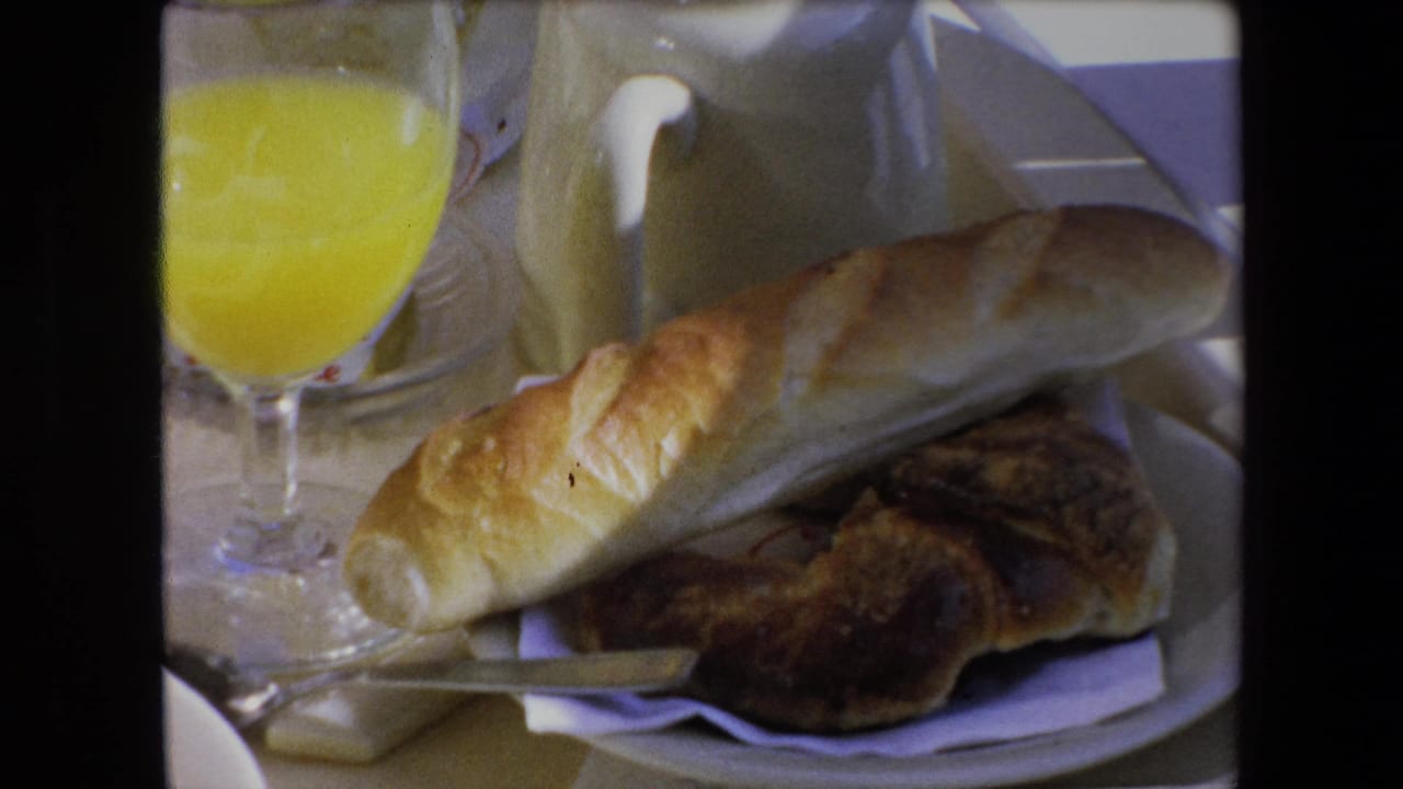 1971 Algeria 8mm frame showing a close tabletop breakfast with a baguette, flaky pastry, white napkin, plate, and bright orange juice.