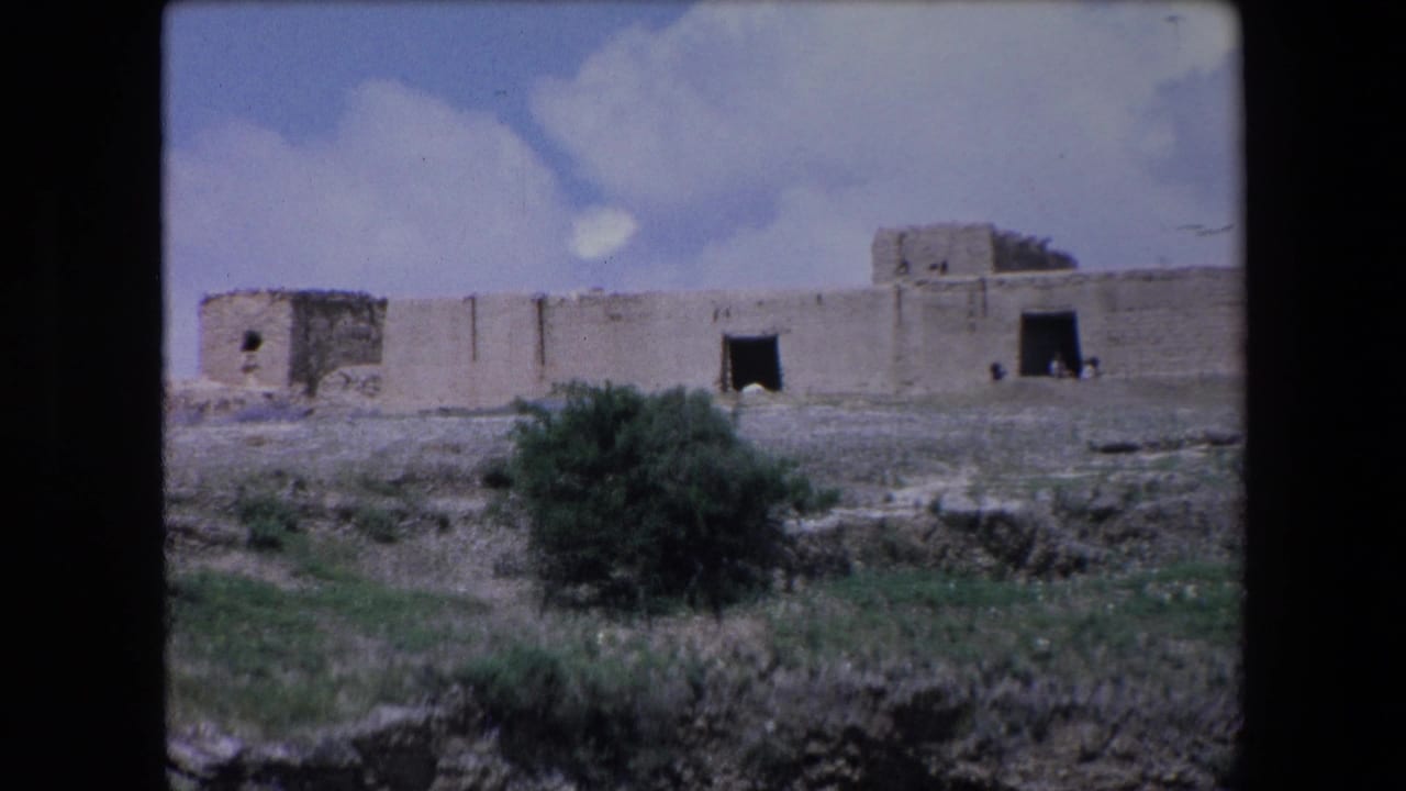 1971 Algeria 8mm frame showing a weathered stone building or fort-like structure across sparse ground under a blue, clouded sky.