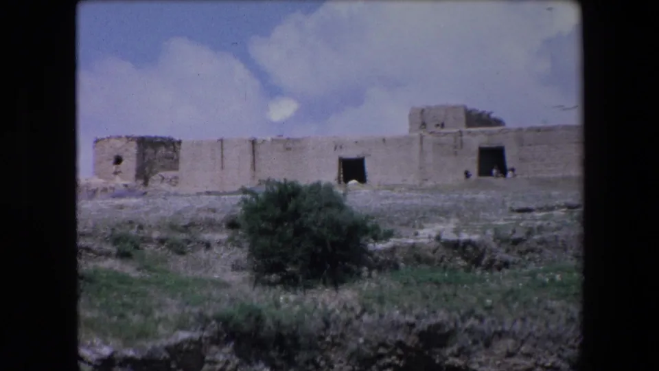 1971 Algeria 8mm frame showing a weathered stone building or fort-like structure across sparse ground under a blue, clouded sky.