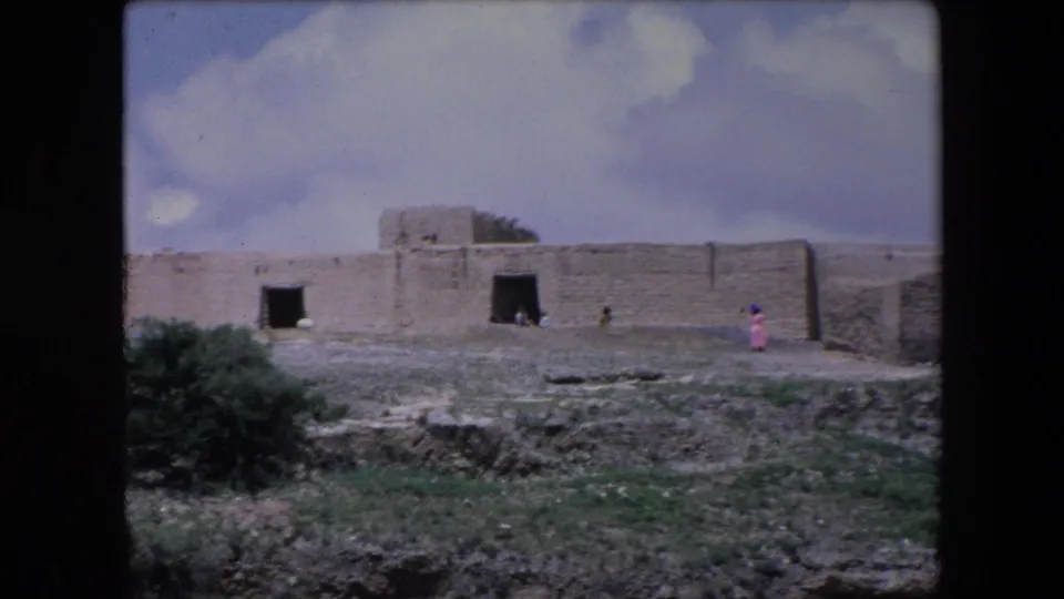 1971 Algeria 8mm frame showing a wide view of a weathered stone structure with sparse greenery and a small person in pink walking across the foreground area.
