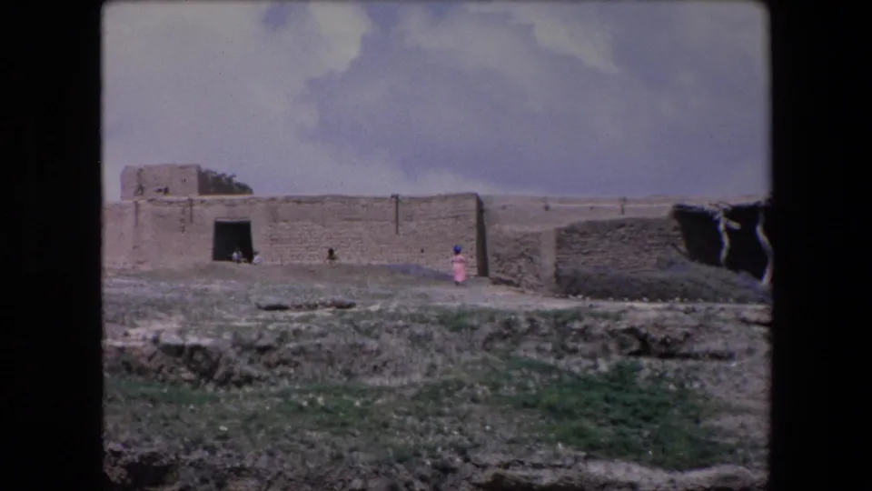 1971 Algeria 8mm frame showing a distant person in pink walking beside a long weathered stone wall and building in a dry, open landscape.