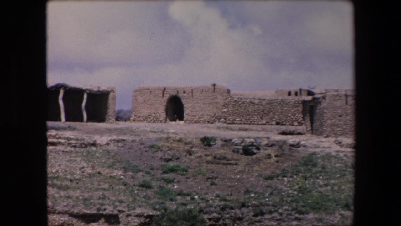 1971 Algeria 8mm frame showing a remote mud-brick or adobe walled structure with an arched doorway, low stonework, sparse shrubs, and cloudy sky.