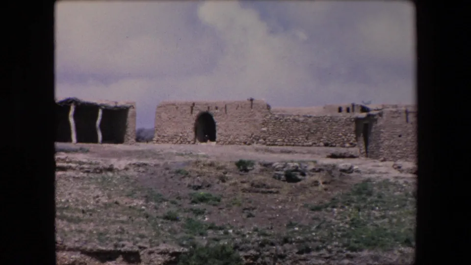 1971 Algeria 8mm frame showing a remote mud-brick or adobe walled structure with an arched doorway, low stonework, sparse shrubs, and cloudy sky.