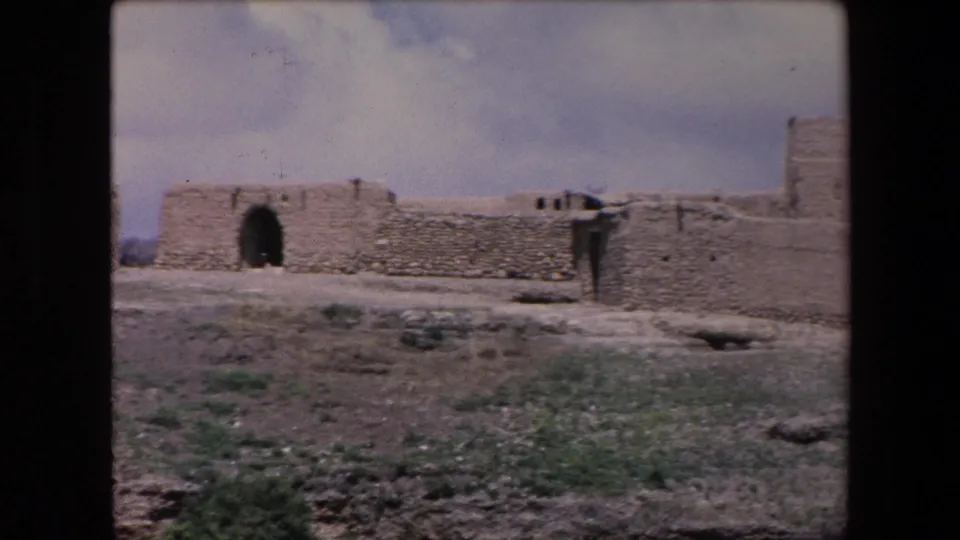 1971 Algeria 8mm frame of a sun-baked walled structure with a dark arched opening, weathered stone or adobe surfaces, dry foreground, and scattered low vegetation.