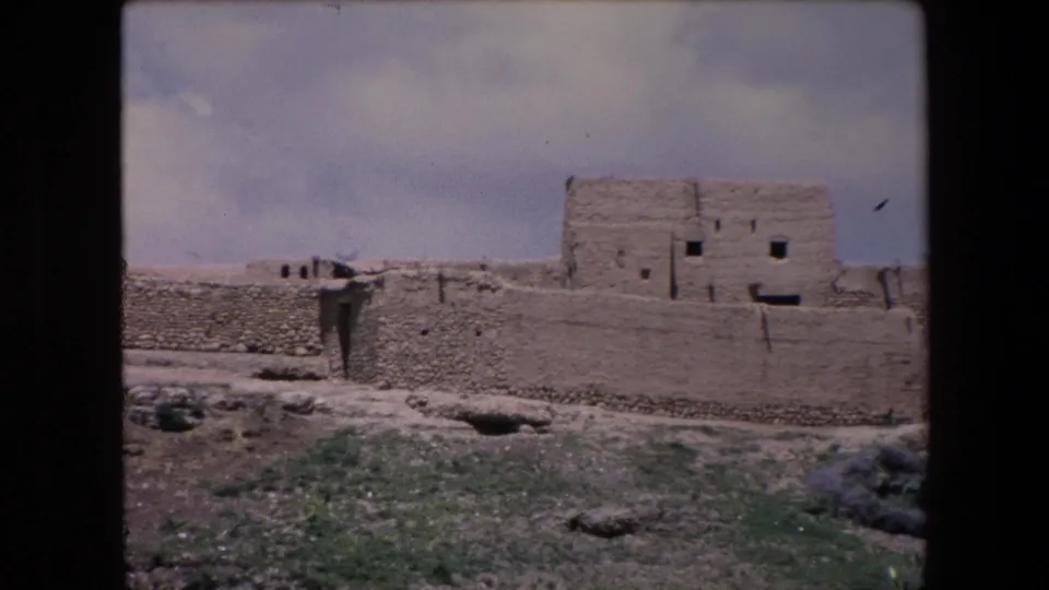 1971 Algeria 8mm frame showing the camera pan reaching a taller adobe building section with small square window openings, a long outer wall, scrub, and rocky desert terrain.