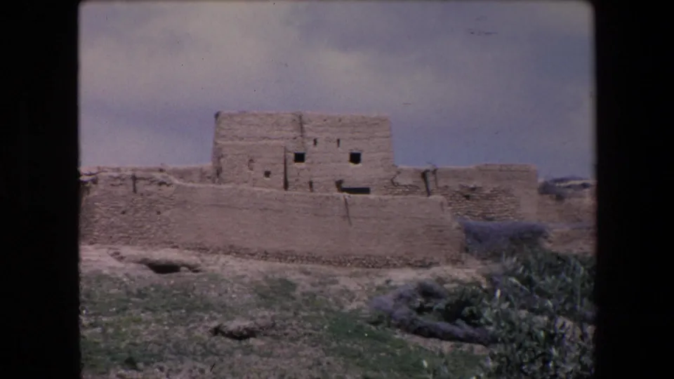 1971 Algeria 8mm frame of a remote mud-brick walled structure with a taller central block, small dark openings, rough foreground earth, and a shrub at the lower right edge.