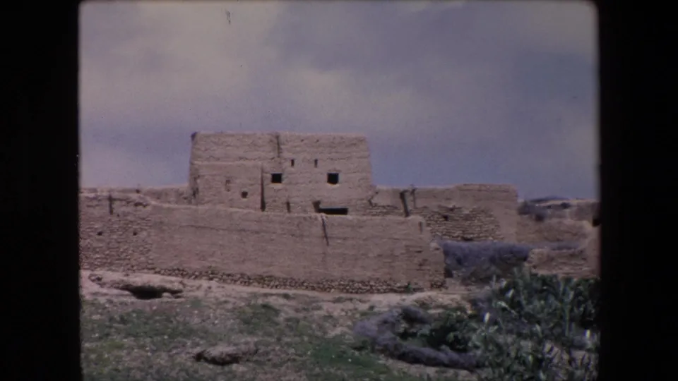 1971 Algeria 8mm frame showing weathered adobe or mud-brick village buildings seen from a distance across sparse green shrubs and dry ground.