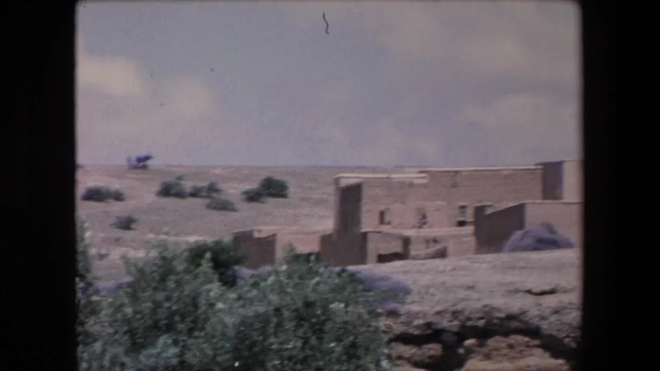 1971 Algeria home-movie frame with low earthen buildings beyond foreground shrubs, open dry terrain, and a few very distant human figures near the structures.